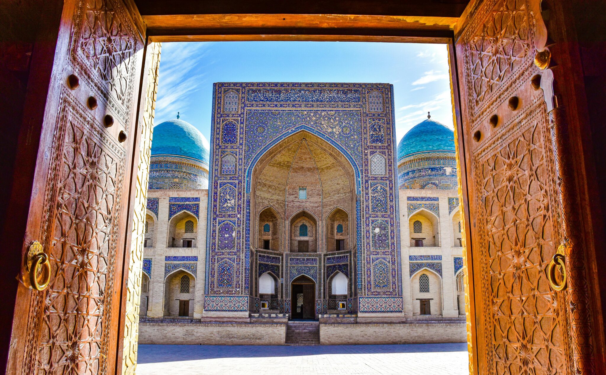 Stunning view of an ornate Uzbek madrasah with blue domes through a carved doorway.